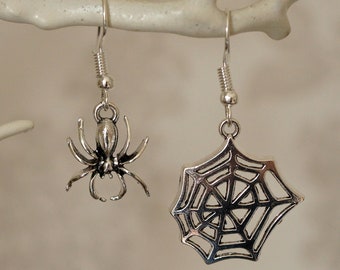 May include: A pair of silver-colored Halloween earrings. One earring is a spider, and the other is a spider web. The earrings are hanging from a white branch. The spider has eight legs and a detailed body. The web is an open design.