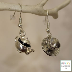 May include: Silver-coloured teapot and teacup earrings. The teapot has a black lid and a curved spout. The teacup sits on a detailed saucer. The earrings are suspended from silver hooks. The earrings are a charming accessory.