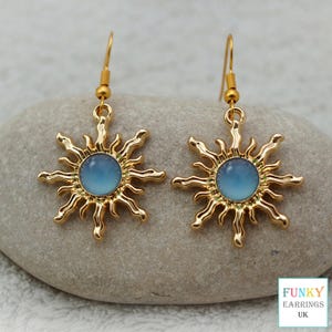 A highly detailed close up of a gold celestial sunburst earrings featuring a smooth blue acrylic centre. The bright gold and soft blue contrast beautifully against the earthy stone background, showcasing this premium everyday boho and astrology accessory.