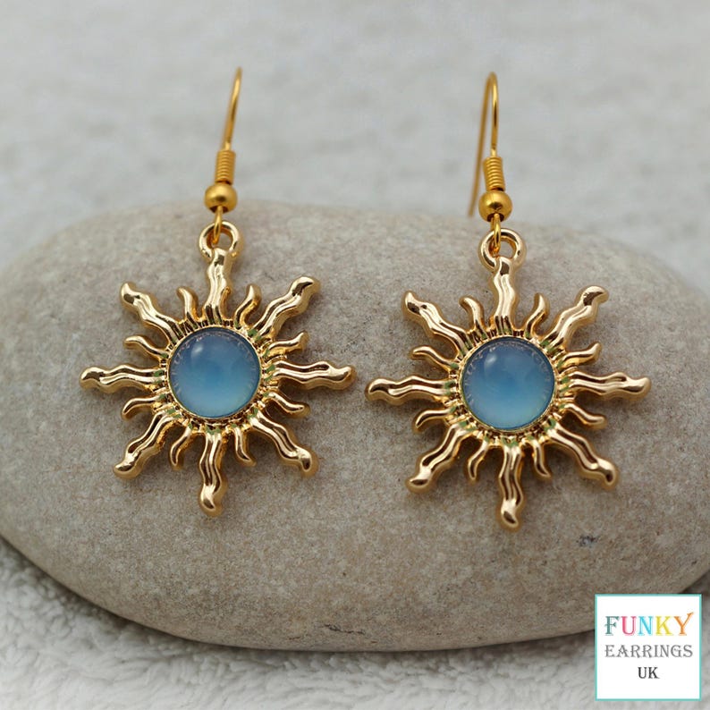 A highly detailed close up of a gold celestial sunburst earrings featuring a smooth blue acrylic centre. The bright gold and soft blue contrast beautifully against the earthy stone background, showcasing this premium everyday boho and astrology accessory.
