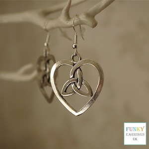 May include: Silver-toned heart-shaped earrings with a Celtic knot design. The earrings are suspended from a fish hook style ear wire. The earrings are displayed against a blurred background, with a white branch in the upper left corner.