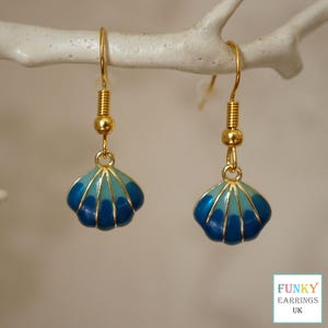 May include: A pair of gold-coloured seashell earrings with blue and light blue enamel detailing. The earrings are hanging from a white branch. The earrings have a gold-coloured hook and a small gold bead.