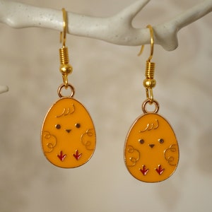 May include: A pair of gold-tone earrings featuring a yellow chick design. The chicks have red feet and black eyes.