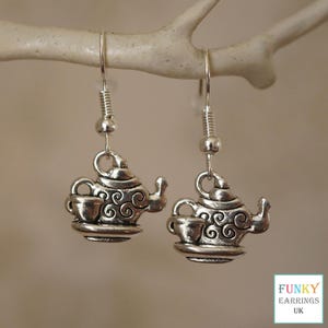 May include: Silver dangle earrings featuring a teapot and cup design. The teapot has a swirl pattern and the cup is on a small plate.