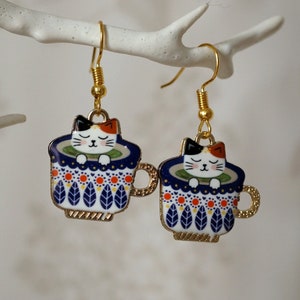 May include: A pair of gold tone earrings featuring a white cat with orange and black markings, sleeping inside a blue and white patterned teacup. The teacup has a gold rim and handle.