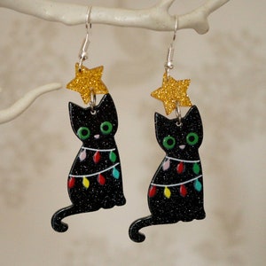 May include: Black cat Christmas tree earrings with a gold glitter star on top. The cat is decorated with colourful lights.