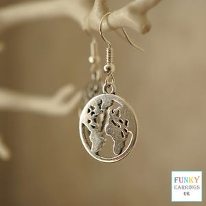May include: Silver-toned dangle earrings featuring a circular pendant with a cut-out design of the Earth's continents. The earrings have a simple hook closure and are displayed against a neutral background. The text "FUNKY EARRINGS UK" is in the bottom right corner.