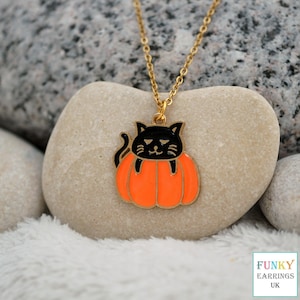 May include: A gold-toned metal necklace featuring an enamel pendant of a black cat emerging from a bright orange pumpkin. The pendant is suspended from a delicate gold-toned chain. The design includes black outlines and details on the cat's face.