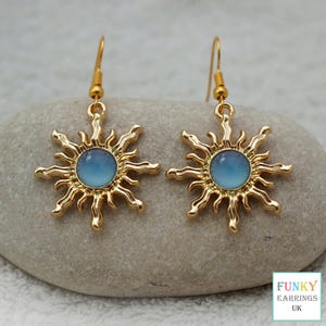 May include: A pair of gold-colored sunburst earrings with a light blue center. The earrings have a hook closure and are displayed on a gray stone. The earrings are from Funky Earrings UK.