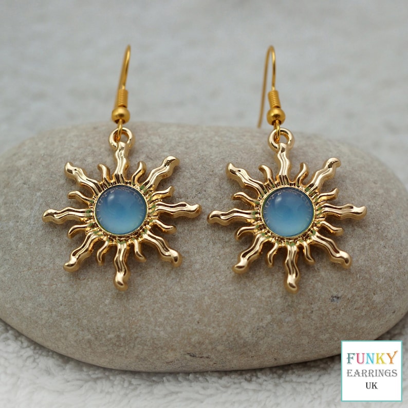 May include: A pair of gold-colored sunburst earrings with a light blue center. The earrings have a hook closure and are displayed on a gray stone. The earrings are from Funky Earrings UK.