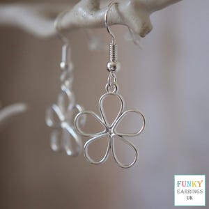 May include: Silver flower earrings with a hook fastening. The earrings are crafted from wire, featuring an open, delicate floral design. They are suspended from a white branch. The text "FUNKY EARRINGS UK" is visible in the bottom right corner.