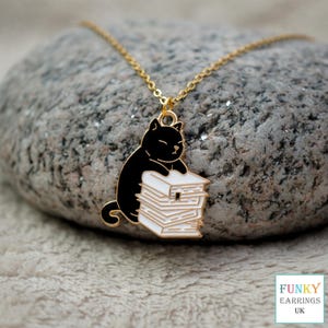 May include: A gold chain necklace with a pendant. The pendant depicts a black cat on a stack of white books. The cat and books have black and white enamel detailing. The necklace is set against a textured grey stone background.