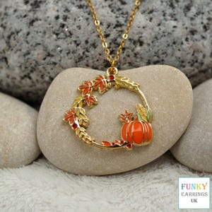 May include: A gold-tone necklace featuring a circular pendant shaped like an autumn wreath. The pendant is decorated with orange and red enamel maple leaves, wheat stalks, and a bright orange pumpkin. The chain is a delicate gold-tone metal link.