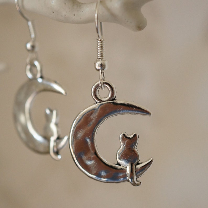 May include: Silver earrings with a crescent moon design and a small cat sitting on the moon.