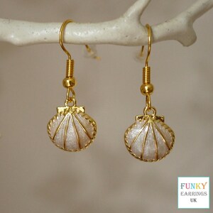 May include: A pair of gold-coloured seashell earrings. Each earring features a pearlescent, fan-shaped shell pendant with gold-coloured trim, suspended from a gold-coloured hook. The earrings are displayed on a white branch.