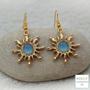 A close up, straight on view of gold blue sunburst drop earrings resting on a smooth, natural grey stone. The celestial earrings feature wavy gold rays surrounding a smooth, blue acrylic centre, neatly arranged to showcase this trendy earthy boho jewellery design.