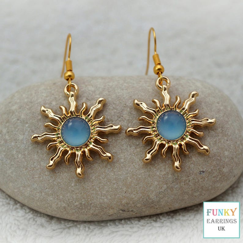 A close up, straight on view of gold blue sunburst drop earrings resting on a smooth, natural grey stone. The celestial earrings feature wavy gold rays surrounding a smooth, blue acrylic centre, neatly arranged to showcase this trendy earthy boho jewellery design.