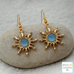 May include: A pair of gold-colored sunburst earrings with a light blue center. The earrings have a hook closure and are displayed on a gray stone. The text "FUNKY EARRINGS UK" is in a white box in the lower right corner.