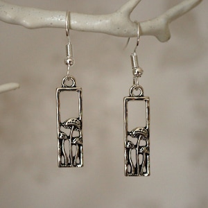 May include: Silver-toned dangle earrings featuring a rectangular frame design with a cluster of mushroom silhouettes. The earrings have a hook closure and are suspended from a small silver ball.