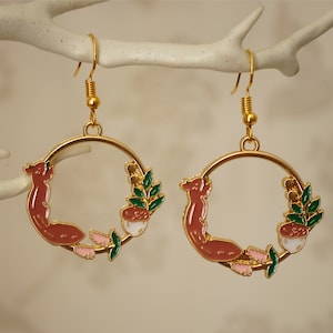 May include: A pair of gold-colored hoop earrings. Each earring features a brown squirrel, green leaves, and a white and brown acorn design. The earrings are suspended from gold-colored ear wires.