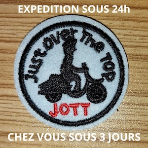 Peut inclure: Un écusson circulaire avec un fond bleu clair et une bordure noire. L'écusson présente un graphisme noir d'une personne conduisant un scooter, avec le texte "Just over the top" en cercle. Le mot "JOTT" est en rouge.