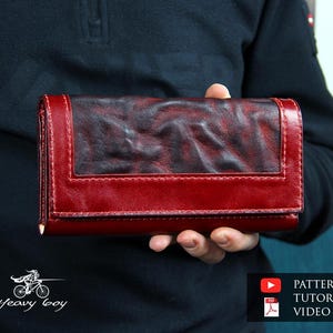 May include: A red leather wallet with a dark, textured center panel. The wallet is rectangular with a flap closure and red stitching. The image also includes a logo and text that says "PATTERN & TUTORIAL VIDEO".