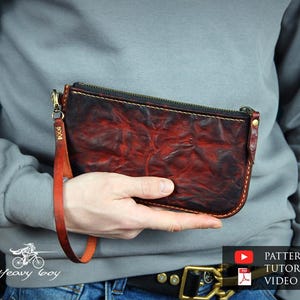 May include: A handcrafted leather clutch in a rich, distressed burgundy color. The rectangular pouch features a black zipper, a wrist strap, and a gold-toned metal clasp. The leather has a textured, mottled appearance.
