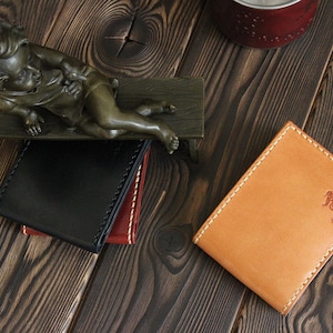 May include: Two leather wallets on a wooden surface. One wallet is black with a brown stitch detail. The other wallet is tan with a brown stitch detail and the text "Crazy Easy" embossed on the front.