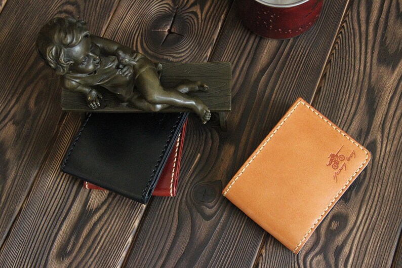 May include: Two leather wallets on a wooden surface. One wallet is black with a brown stitch detail. The other wallet is tan with a brown stitch detail and the text "Crazy Easy" embossed on the front.