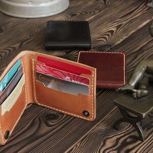 May include: A brown leather wallet with multiple card slots and a snap closure. The wallet is open and has several cards inside. The wallet is on a wooden surface with two other wallets in the background.