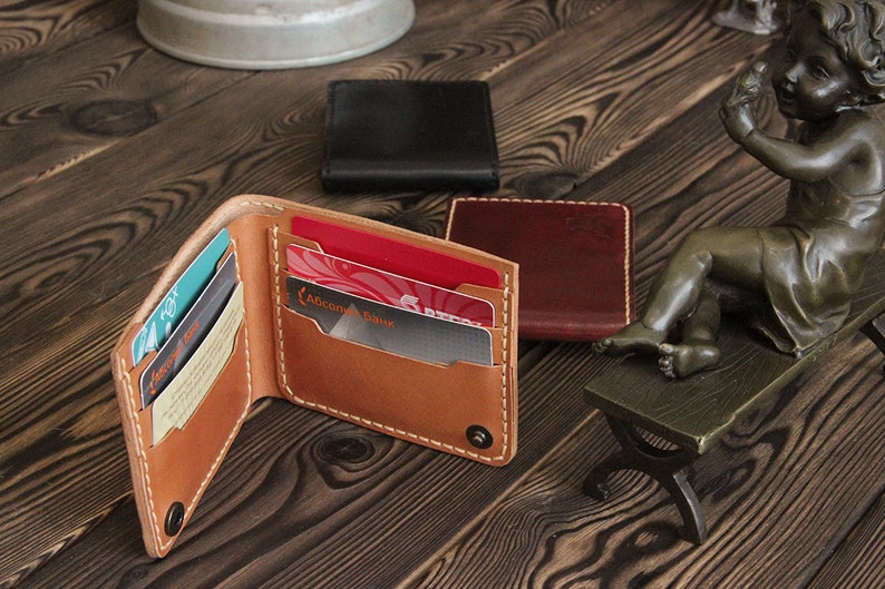May include: A brown leather wallet with multiple card slots and a snap closure. The wallet is open and has several cards inside. The wallet is on a wooden surface with two other wallets in the background.