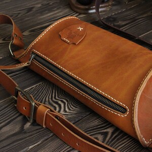 May include: A brown leather crossbody bag with a zipper closure and a small metal emblem. The bag has a long adjustable strap.
