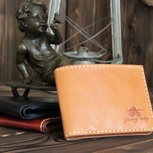 May include: A tan leather wallet with a stitched border and a red logo that says "Heavy Boy" with a motorcycle graphic. The wallet is sitting on a wooden surface.
