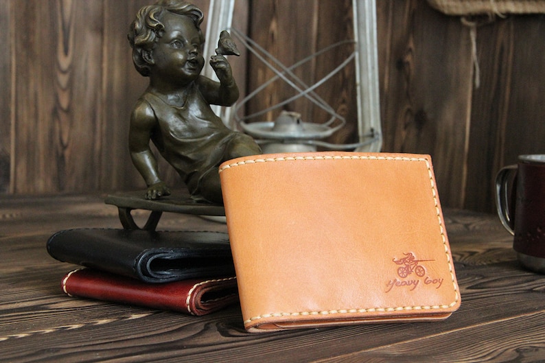 May include: A tan leather wallet with a stitched border and a red logo that says "Heavy Boy" with a motorcycle graphic. The wallet is sitting on a wooden surface.