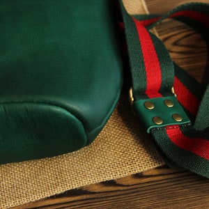 May include: A dark green leather bag with a red and green striped strap. The strap has brass accents and a green leather patch.