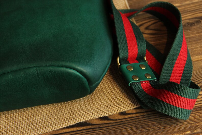 May include: A dark green leather bag with a red and green striped strap. The strap has brass accents and a green leather patch.