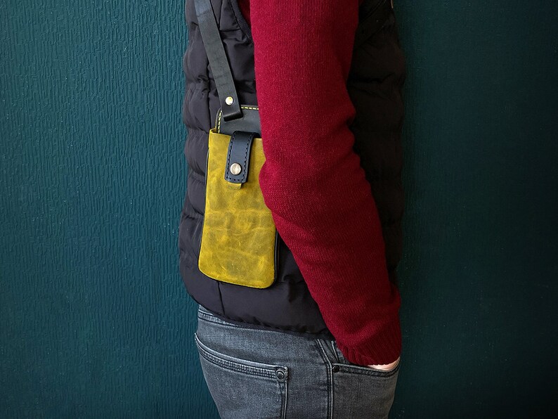 May include: A person wearing a red sweater and a black vest with a yellow leather phone case attached to the vest.