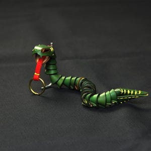 Op de afbeelding: Een groene leren slangen sleutelhanger met een rode tong en een gouden ring. De slang is opgerold en heeft een gedetailleerd ontwerp.