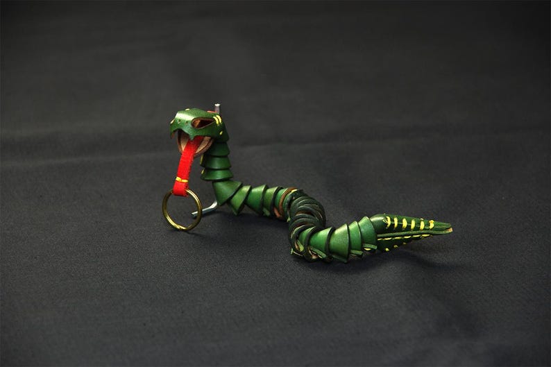 Op de afbeelding: Een groene leren slangen sleutelhanger met een rode tong en een gouden ring. De slang is opgerold en heeft een gedetailleerd ontwerp.
