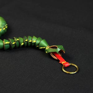 Op de afbeelding: Een groene leren slangen sleutelhanger met gele accenten en een rode en gouden sleutelring.