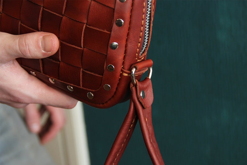 May include: A brown leather bag with a woven design and silver studs. The bag has a zipper closure and a short strap.