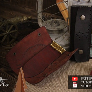 Könnte beinhalten: Zwei Leder-Schlüsselanhänger, einer braun und einer schwarz, mit einem Messing-Schlüsselring. Der braune Schlüsselanhänger hat ein dekoratives Design und der schwarze Schlüsselanhänger hat ein perforiertes Design. Die Schlüsselanhänger liegen auf einer Jutestoff-Oberfläche mit einem kleinen Weihnachtsbaum aus Holz.