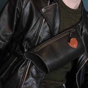 May include: A black leather fanny pack with a brown leather patch and a zipper closure. The fanny pack is worn over a black leather jacket.