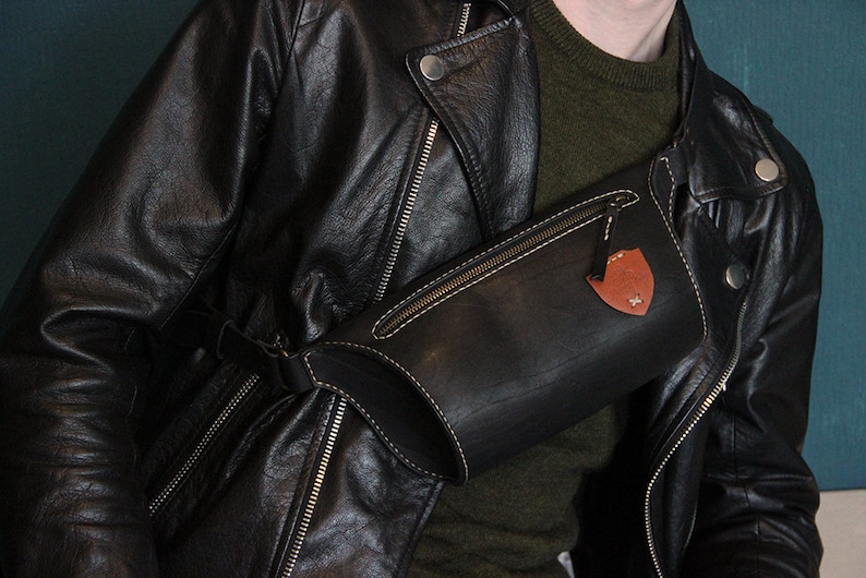 May include: A black leather fanny pack with a brown leather patch and a zipper closure. The fanny pack is worn over a black leather jacket.