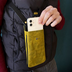 May include: A yellow leather phone case with a black strap attached to a black vest. A white smartphone is inside the case.