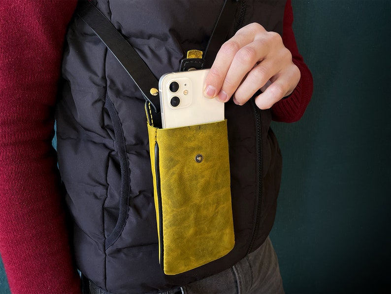 May include: A yellow leather phone case with a black strap attached to a black vest. A white smartphone is inside the case.