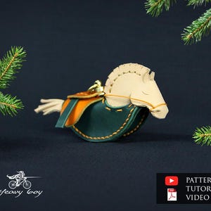 Può includere: Un ornamento a forma di cavallo in pelle fatto a mano, con testa color crema e sella verde e arancione. Il cavallo è ambientato su uno sfondo scuro con rami di sempreverdi, suggerendo un tema festivo. L'ornamento ha un anello dorato per appenderlo.