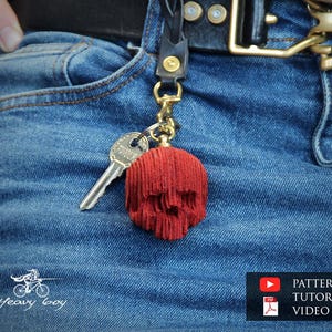以下が含まれることがあります： 赤いスカル型のキーホルダーに鍵が取り付けられ、黒い革ストラップと金色の金具から吊り下げられています。キーホルダーは青いデニムのジーンズに取り付けられています。画像には「PATTERN & TUTORIAL VIDEO」というテキストも含まれています。