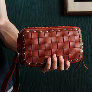 May include: A brown leather clutch with a woven design and gold studs. The clutch has a zipper closure and a strap.