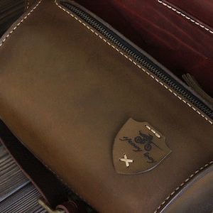 May include: A brown leather bag with a zipper closure and a small leather patch with the text "heavy duty" and a motorcycle graphic. The bag has a strap and is made of high-quality leather.